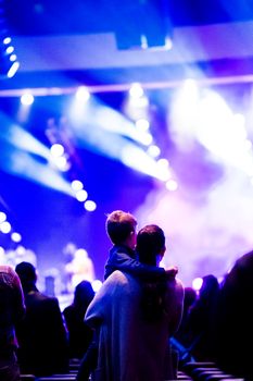A mother carrying her child on her shoulders, enjoying a lively concert with colorful stage lights.