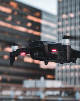 A drone with red lights hovering in an urban city setting at dusk, framed by modern buildings.