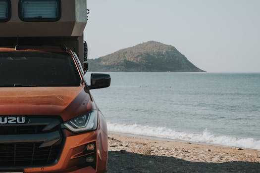 Orange Isuzu pickup truck parked on a beach with scenic sea and mountain view.