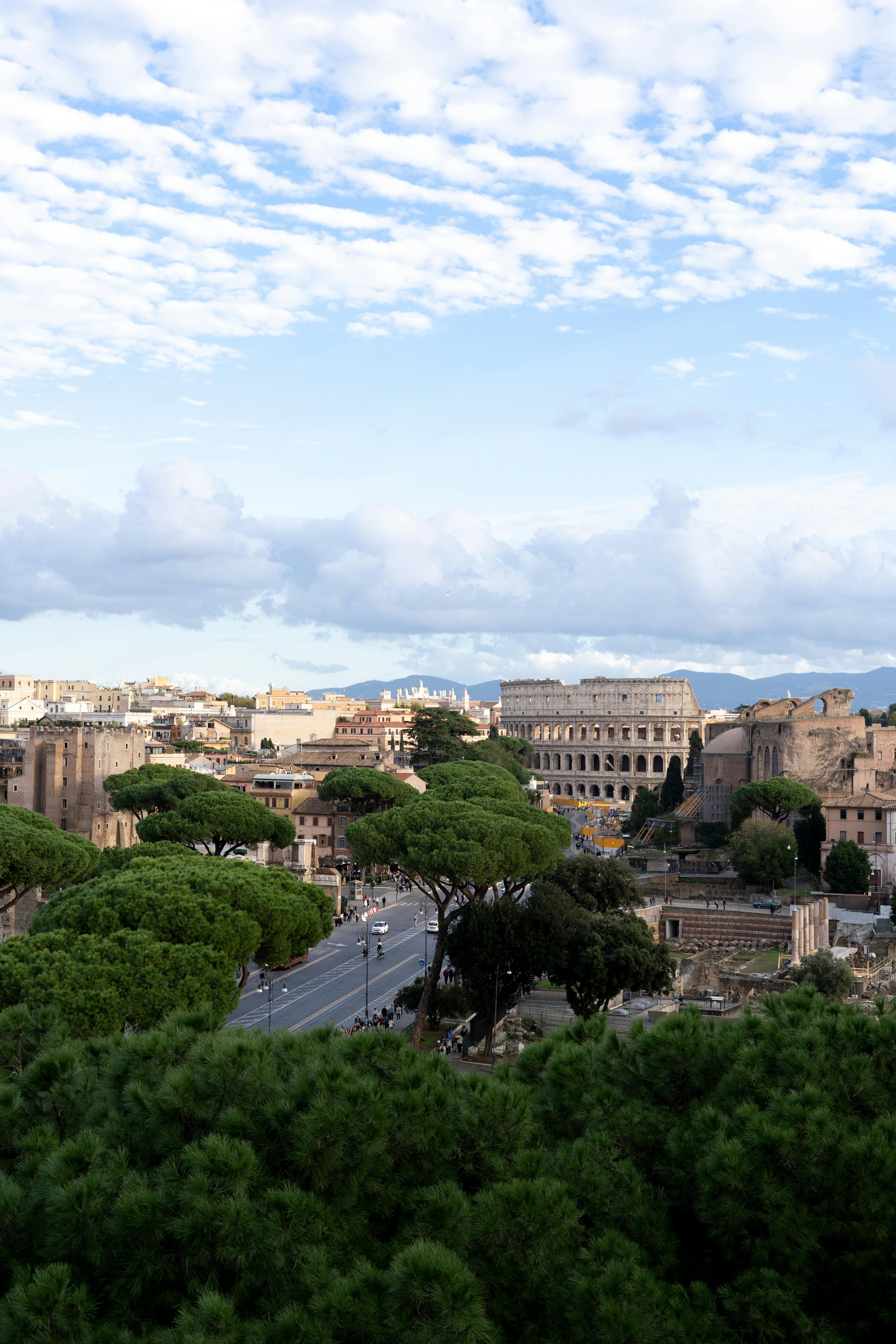 Free Discover the iconic Colosseum amid ancient Roman architecture and lush greenery under a bright sky. Stock Photo