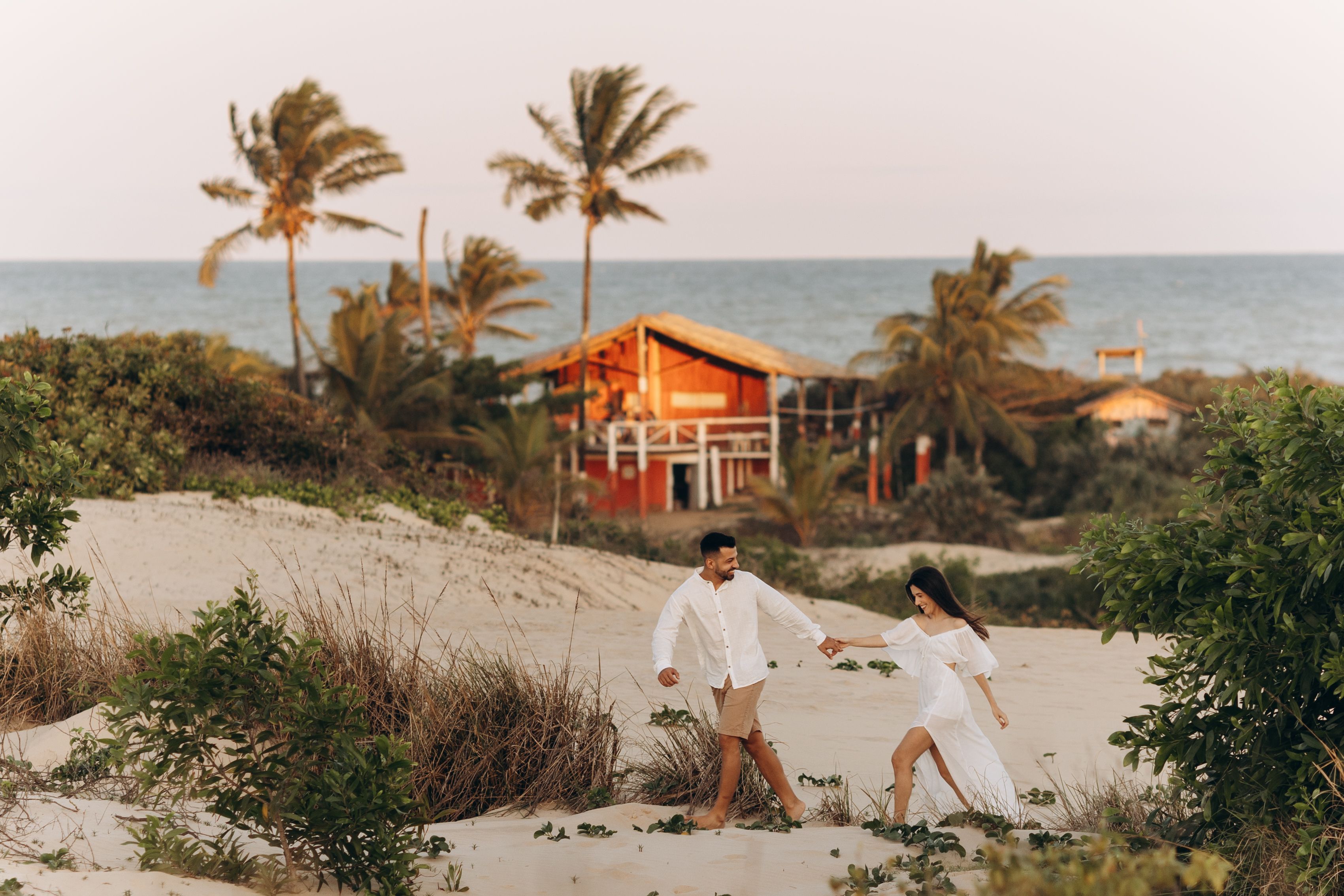 A happy couple walks hand in hand on a sandy beach with a tropical backdrop during sunset, representing love and togetherness.