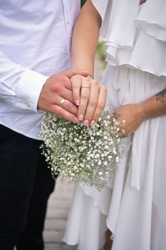Close-up of bride and groom holding hands with a delicate bouquet, showcasing wedding rings.