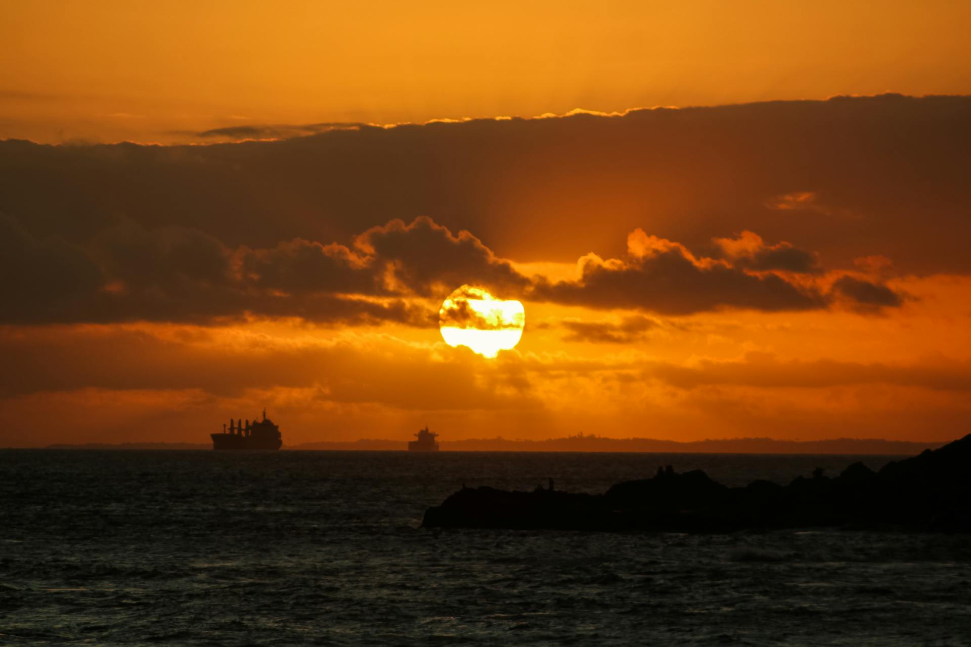 Golden hour sunset over ocean with ships silhouetted against the sky.