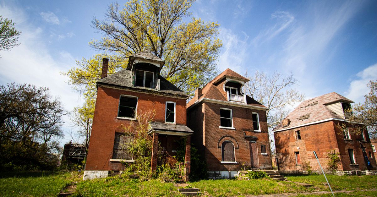 Three abandoned brick houses in a suburban neighborhood under a clear blue sky.