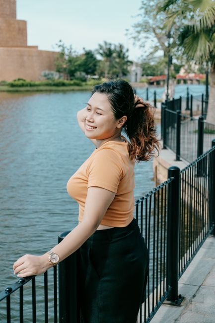 Asian woman smiling by a railing near water in an urban park setting.