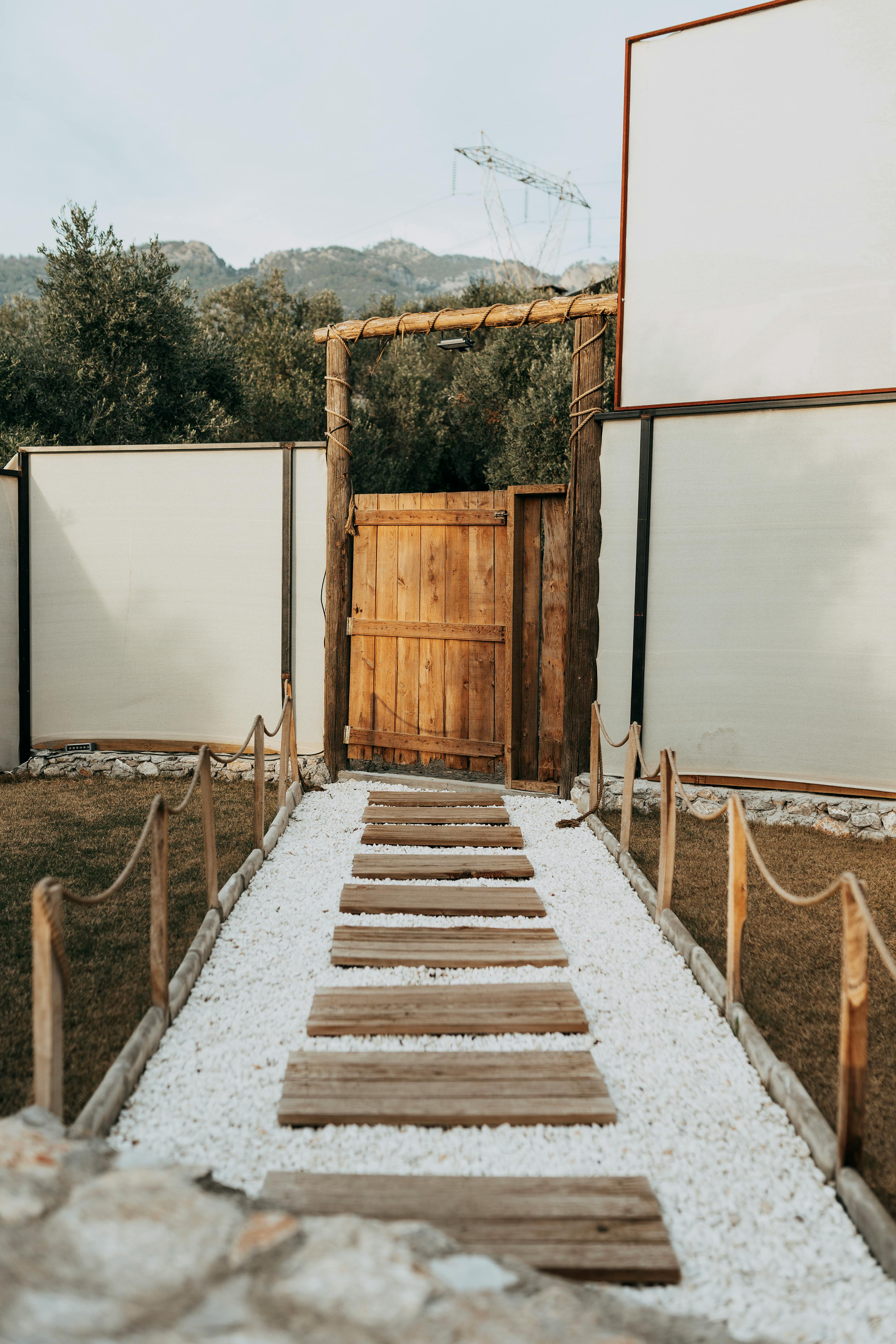 Explore this rustic wooden gate and white pebble path in Ortaca, Muğla, Türkiye, set against a tranquil garden backdrop.