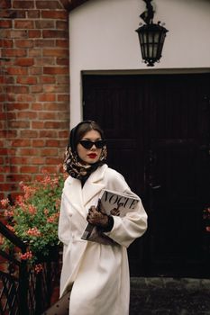 Stylish woman in a coat with headscarf holding a fashion magazine, exuding classic glamour.