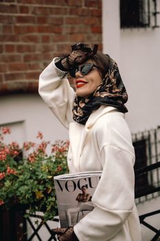 Fashionable woman with red lips and headscarf holding a Vogue magazine outdoors.