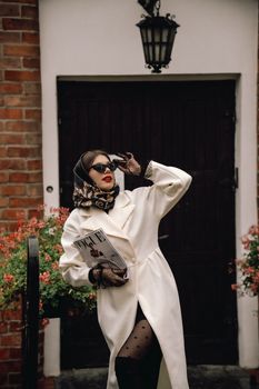 Stylish woman poses in white coat with sunglasses, scarf, and magazine in outdoor setting.