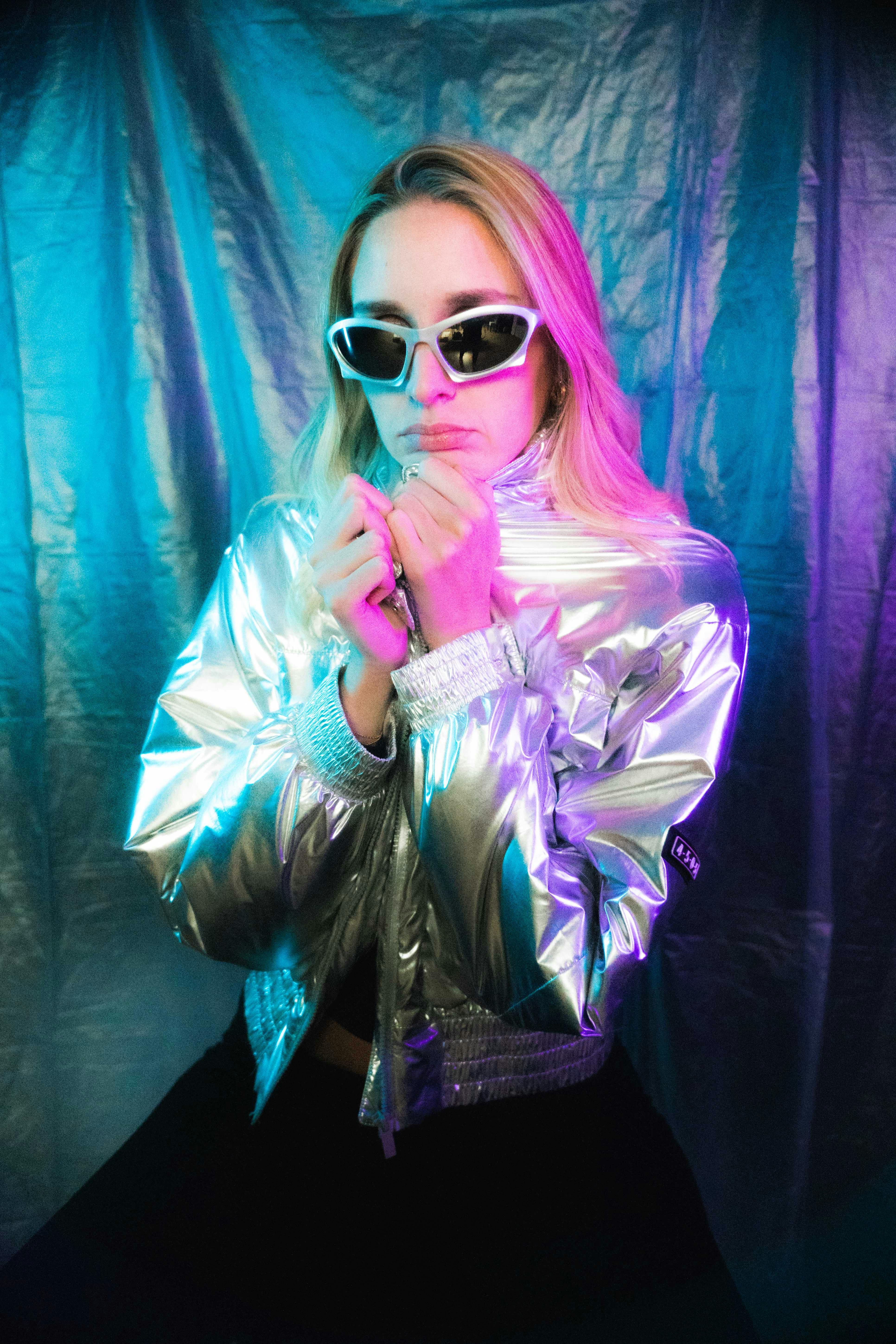 Stylish woman in shiny silver jacket and sunglasses poses against vibrant backdrop.