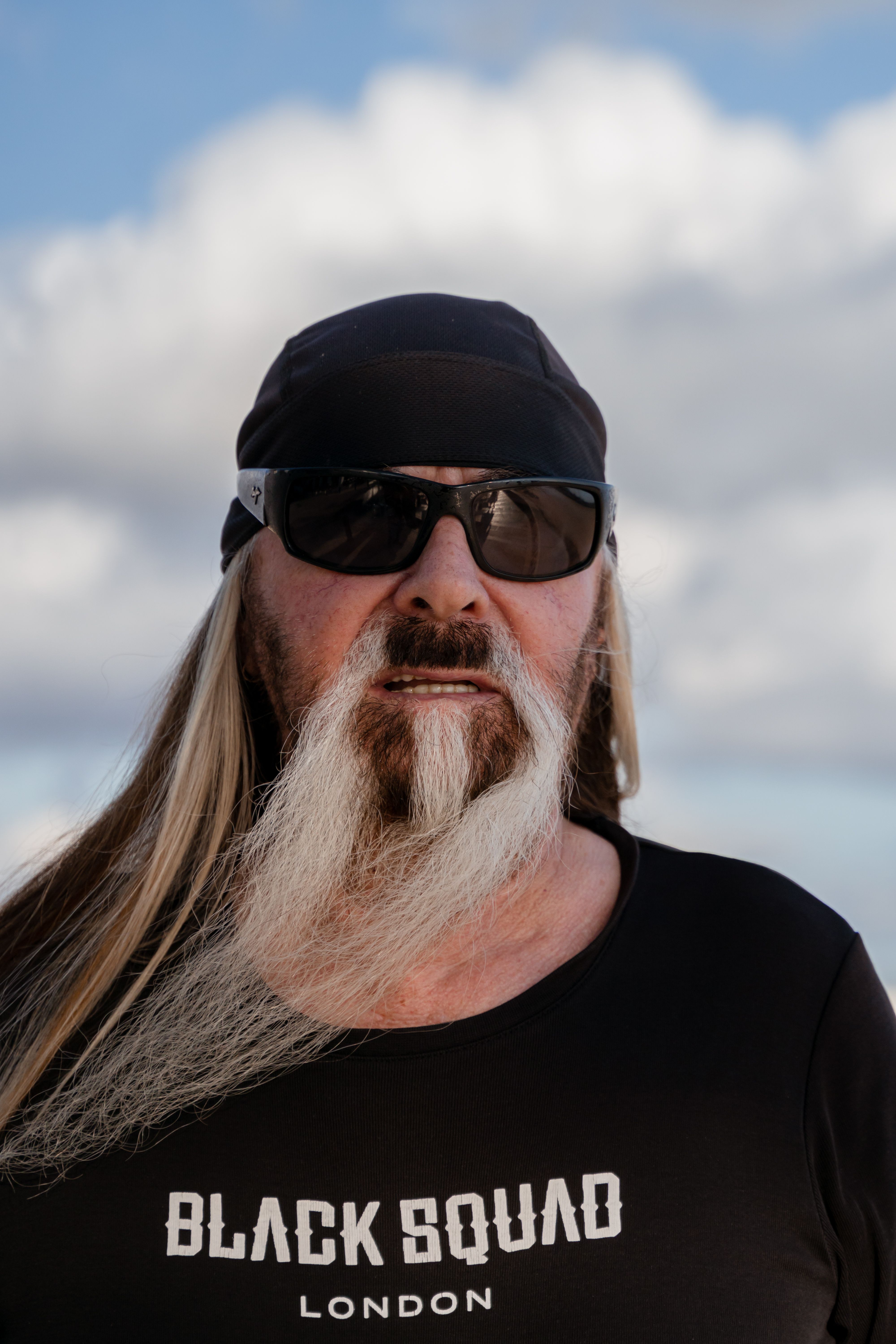 Portrait of an elderly man with long beard, sunglasses, and cap under a cloudy sky.