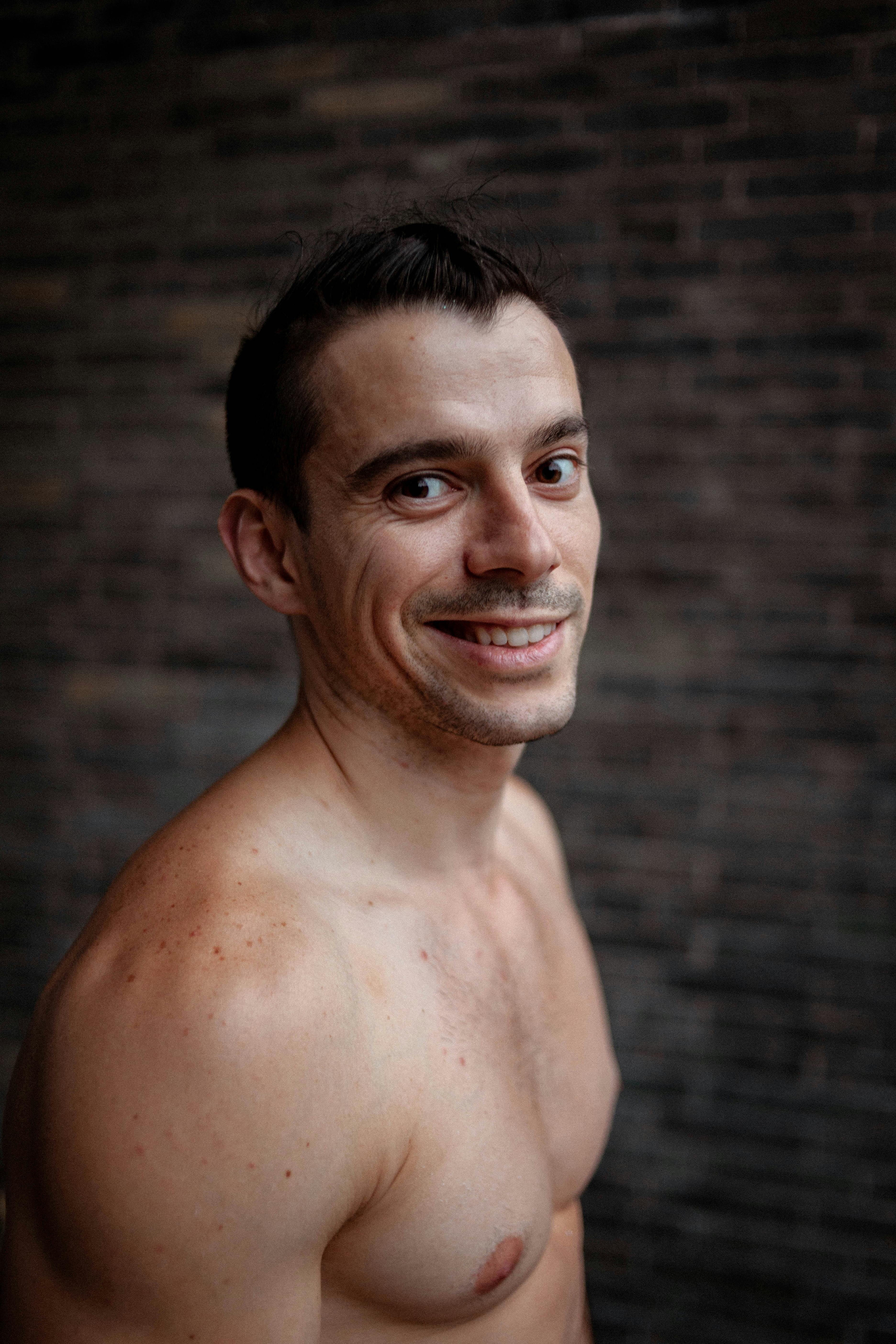 Portrait of a smiling shirtless man with muscular build against a brick wall.