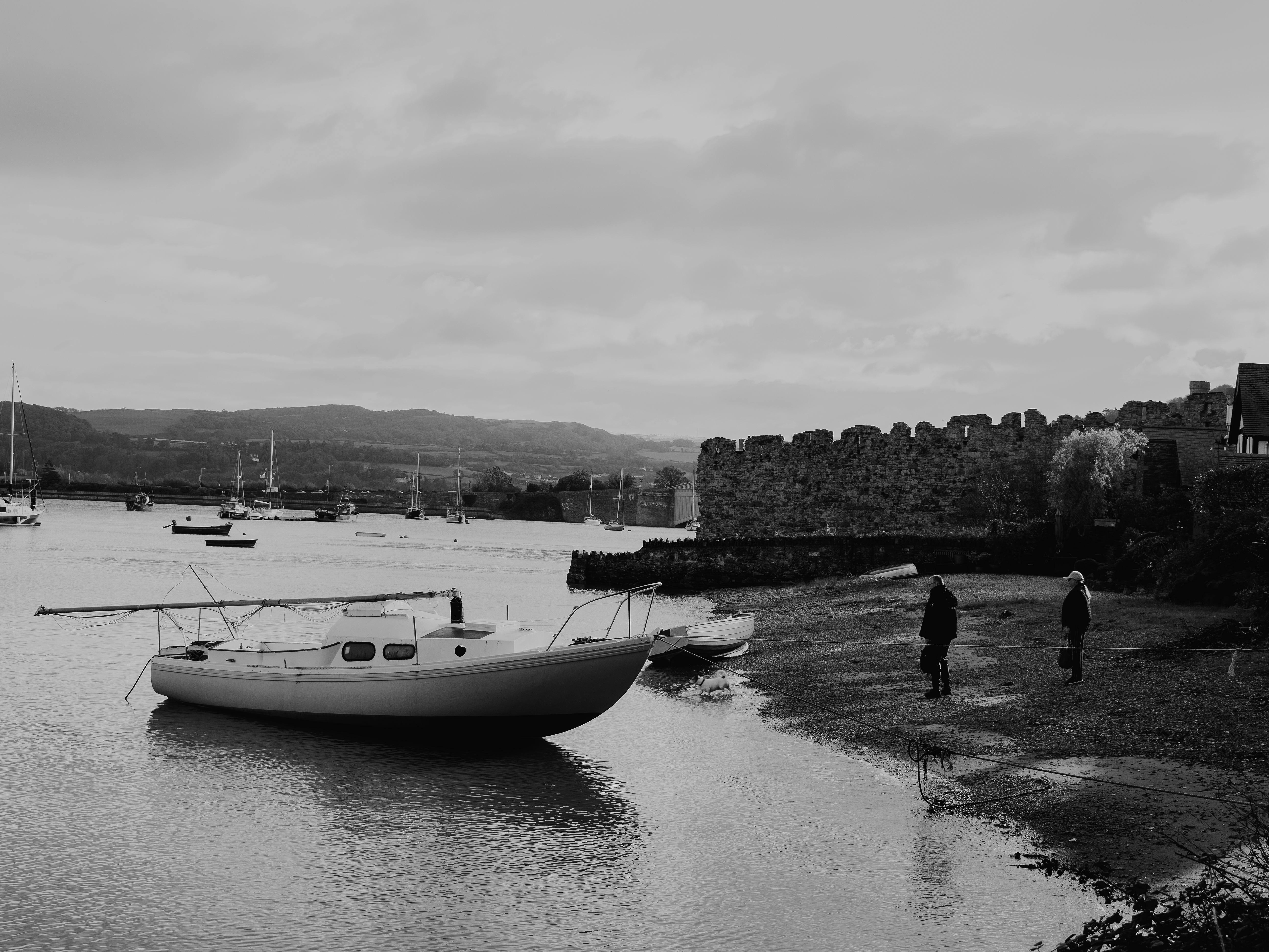 Black and white photo of moored boats by Conwy Castle, Wales, with coastal scenery.