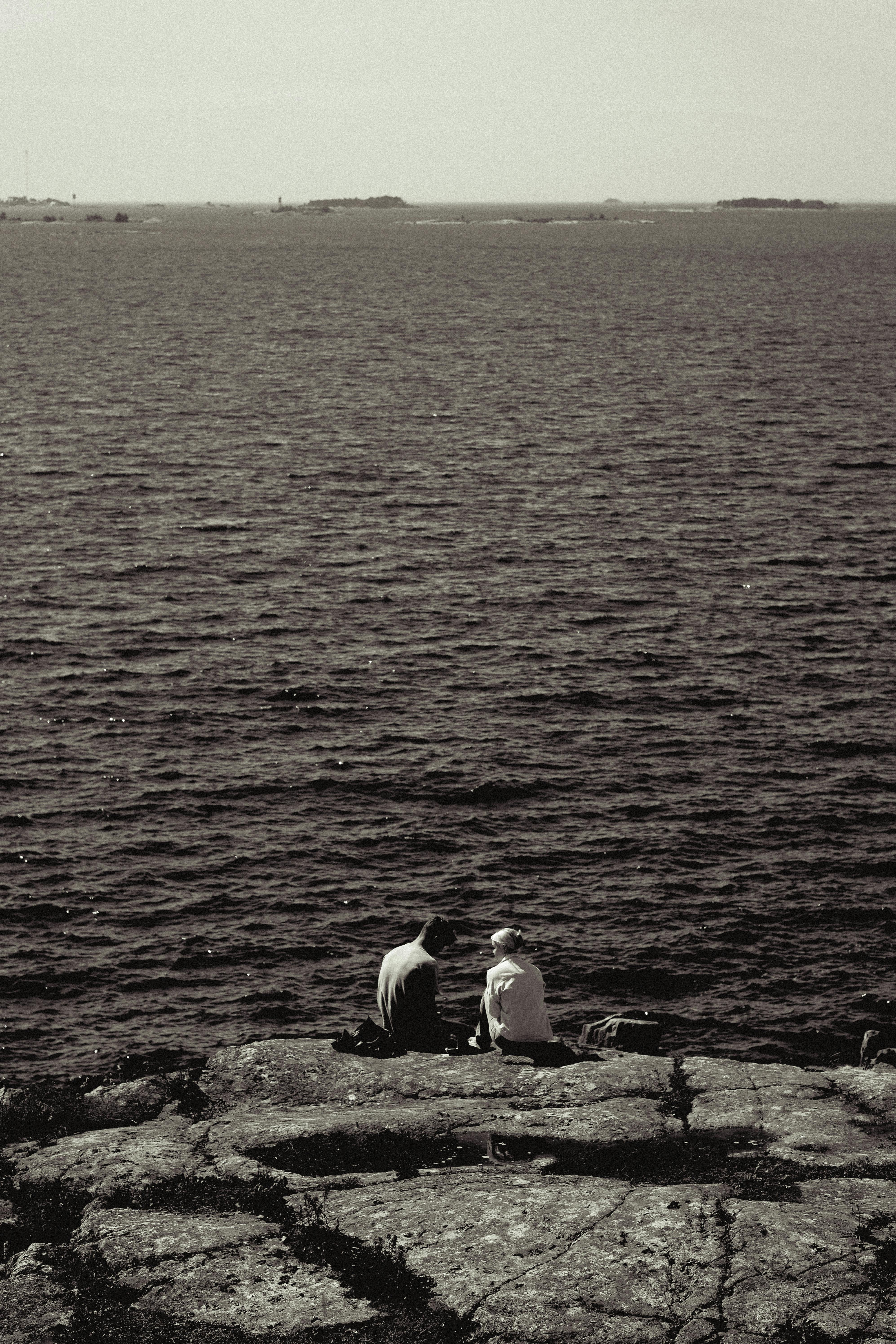Two people sitting on rocky shore in Helsinki, Finland overlooking the sea. Monochrome photo.