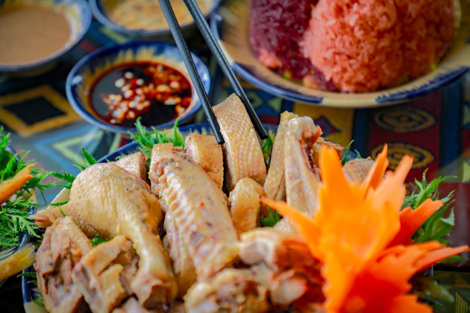 A whole glossy, dark mahogany soy sauce poached chicken chopped into pieces and arranged on a platter, garnished with scallions and a small bowl of reserved sauce alongside steamed rice.