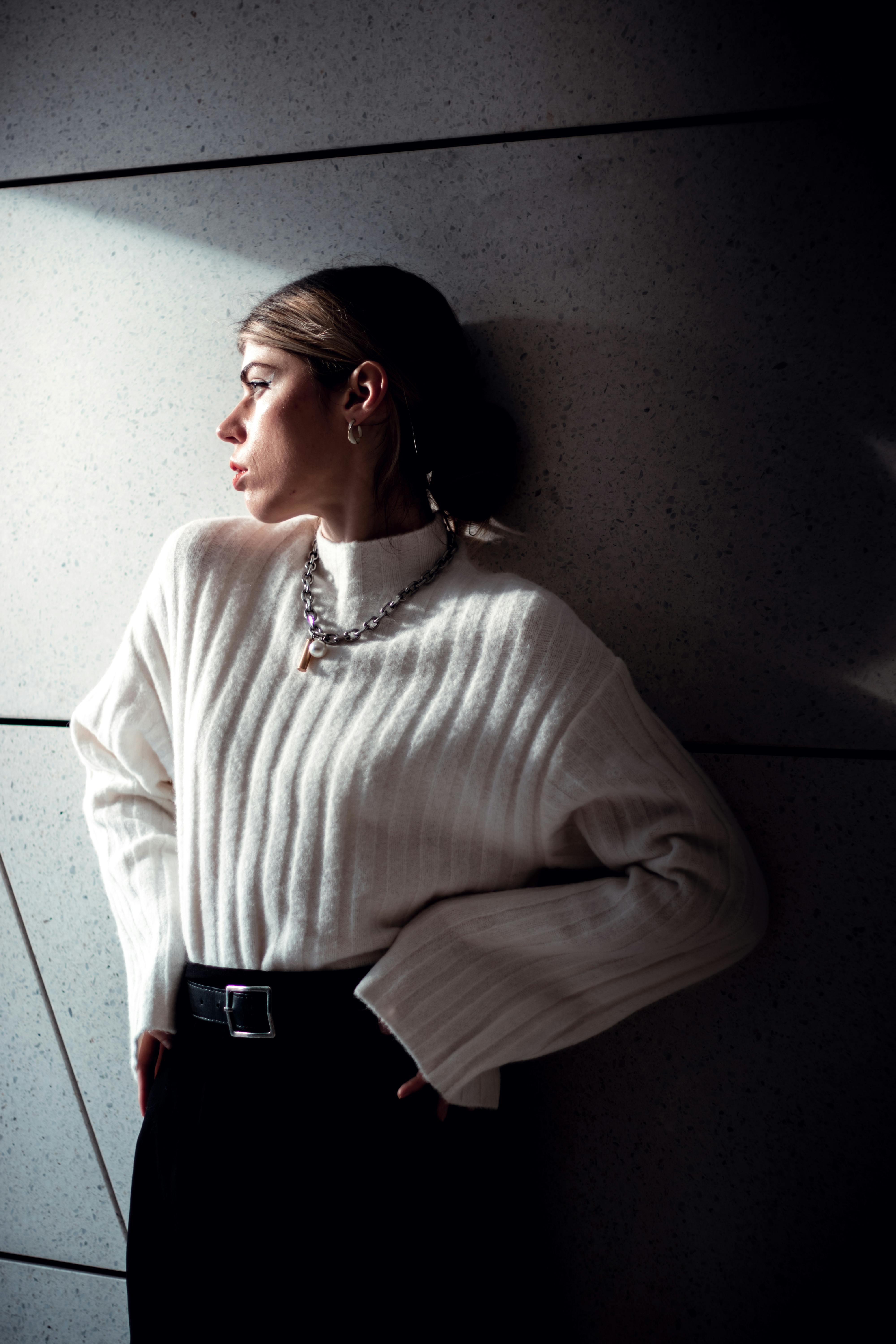 Fashionable woman in a white sweater posing against a modern wall with dramatic lighting.