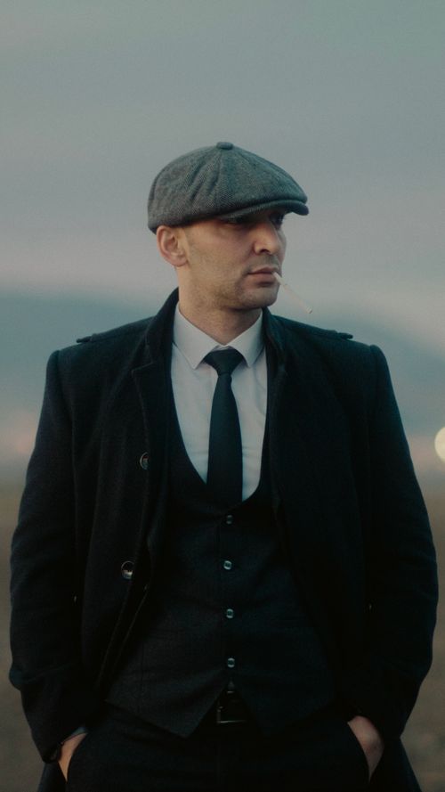 Free A stylish man wearing a suit and flat cap, standing outdoors with a serene backdrop, exuding elegance and confidence. Stock Photo