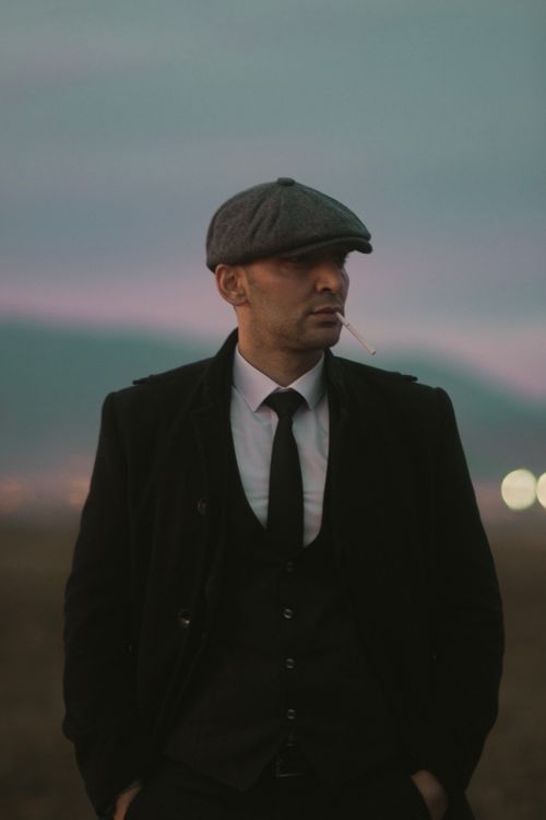 Free Portrait of a stylish man in a suit and flat cap smoking a cigarette outdoors at dusk. Stock Photo