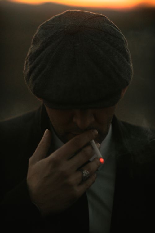 Free Dramatic low-light portrait of a man smoking with a flat cap, evoking vintage style. Stock Photo