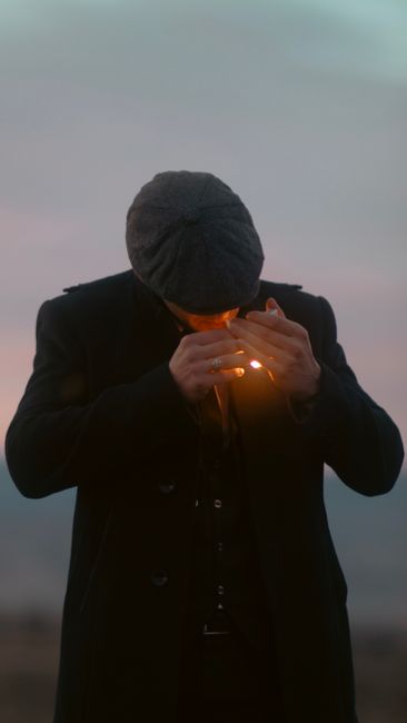 A man in a suit and hat lights a cigarette outdoors during twilight.
