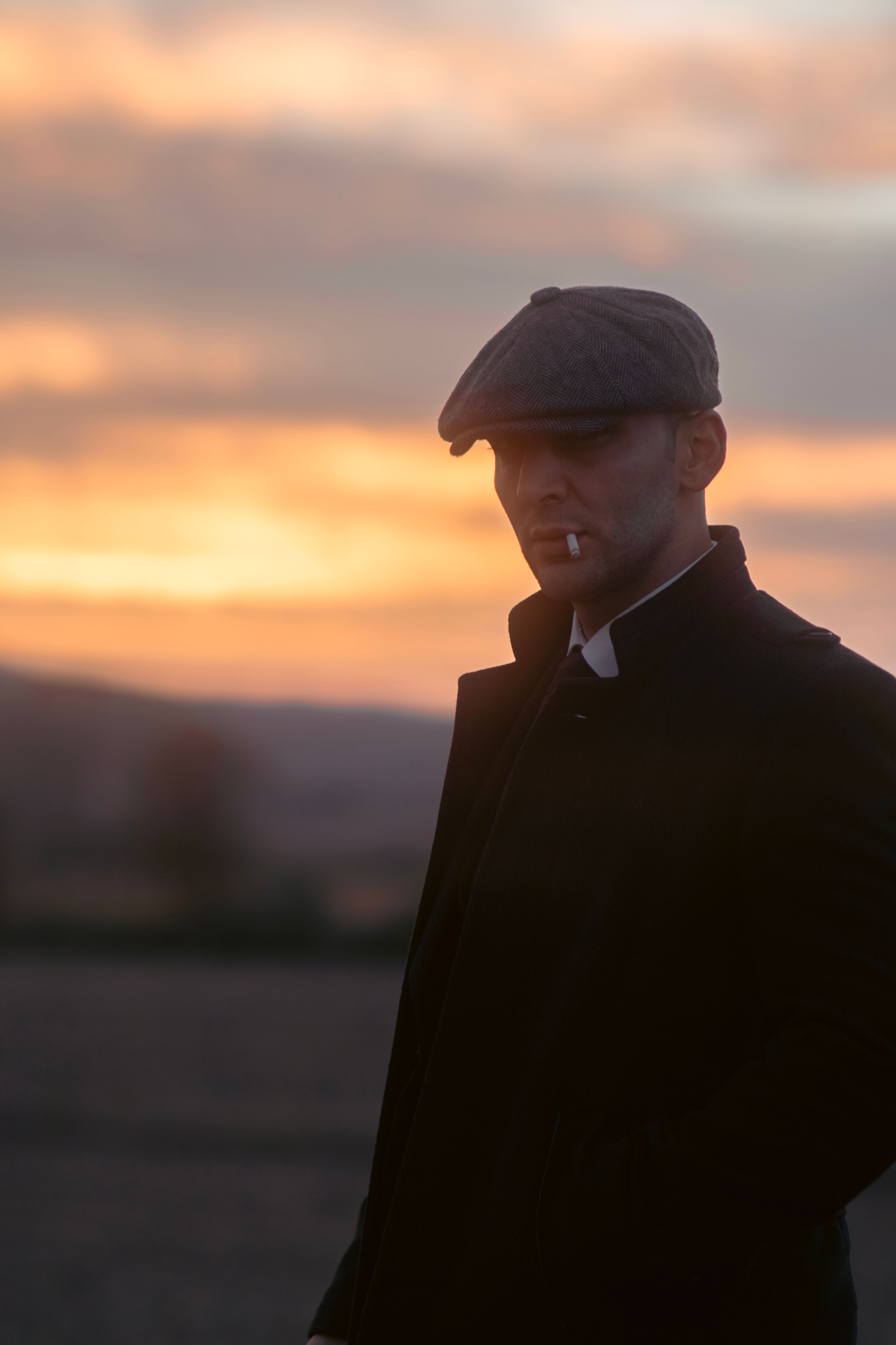 A man wearing a hat and jacket smokes a cigarette during a dramatic sunset outdoors.