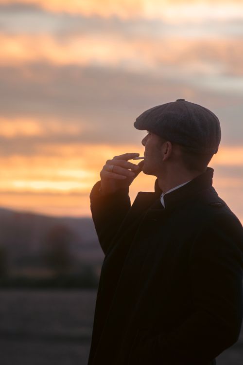 Free A fashionable man in a peaked cap smoking outdoors at sunset, evoking a vintage style. Stock Photo