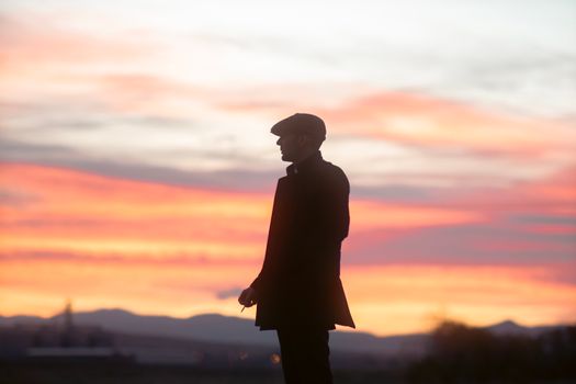 Silhouette of a man wearing a hat and jacket standing in the countryside at sunset.
