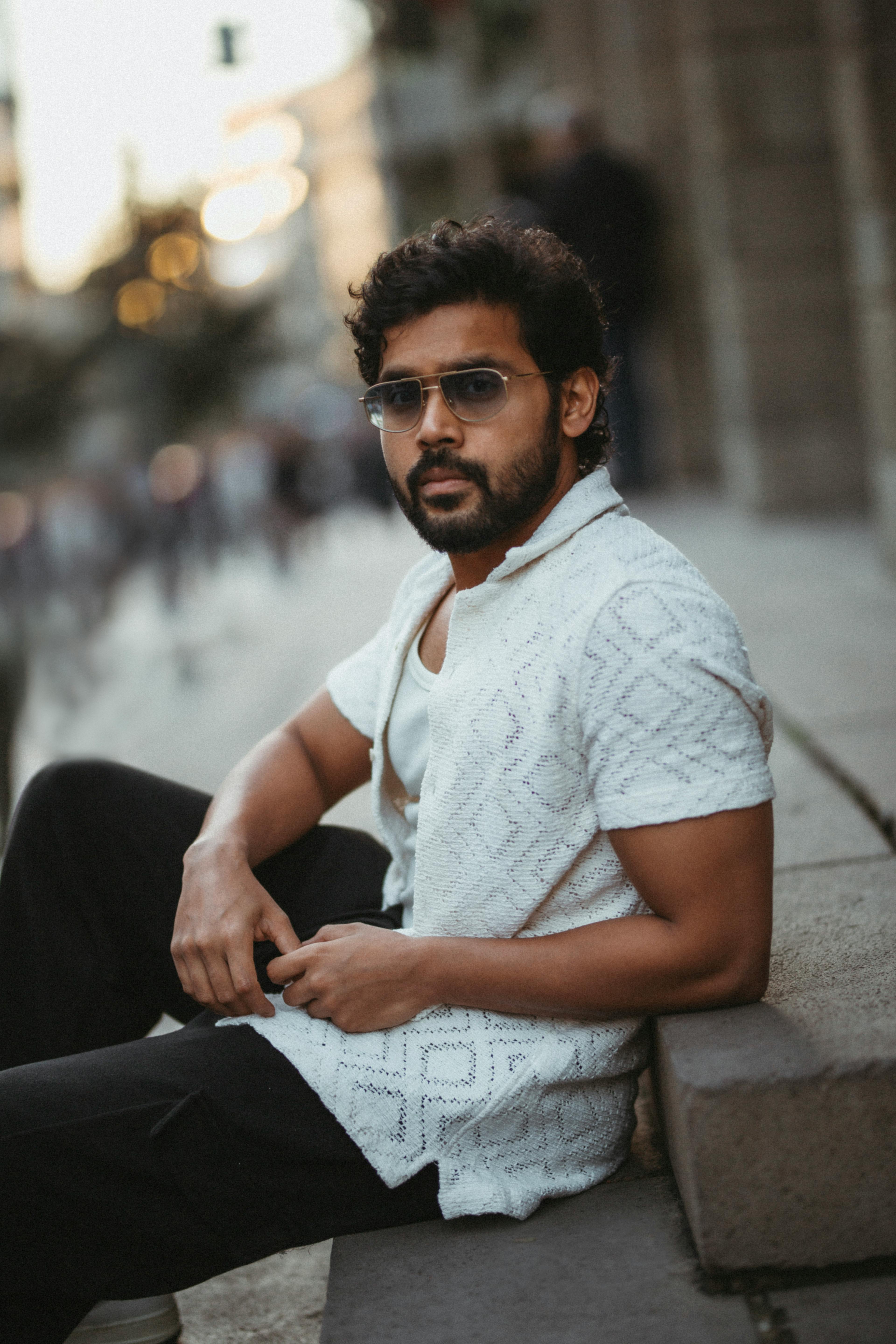 Portrait of a stylish man with sunglasses sitting on city stairs at dusk, exuding elegance.