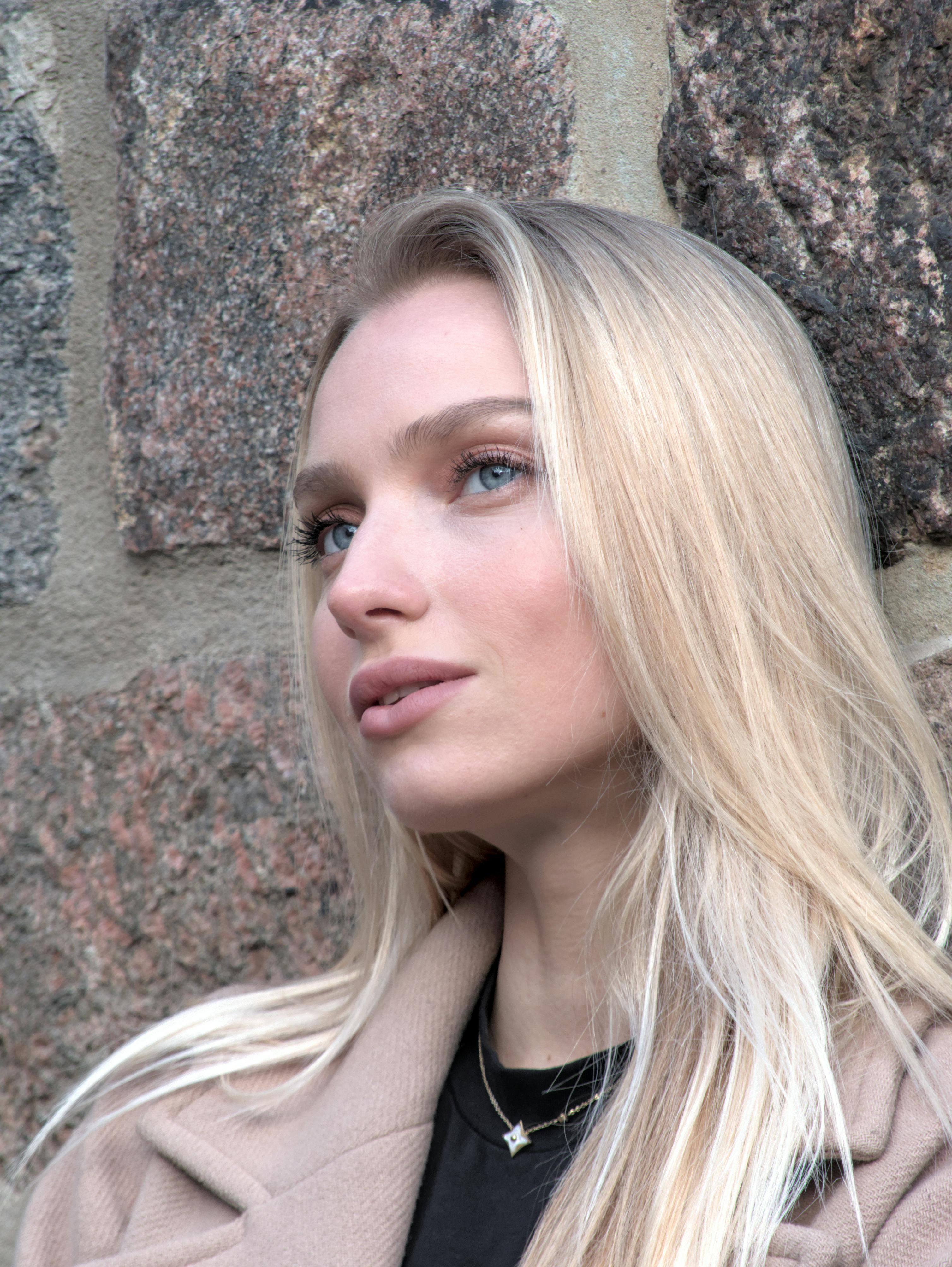 Close-up portrait of a young woman with blonde hair and blue eyes against a stone wall.