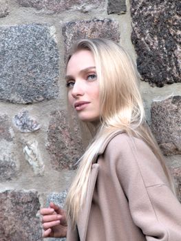 Stylish young woman with a beige coat poses elegantly against a rustic stone wall in Berlin.
