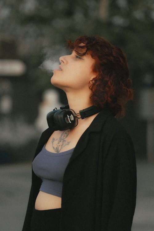 Young Woman with Headphones Standing Outside and Releasing Smoke out of Her Mouth 