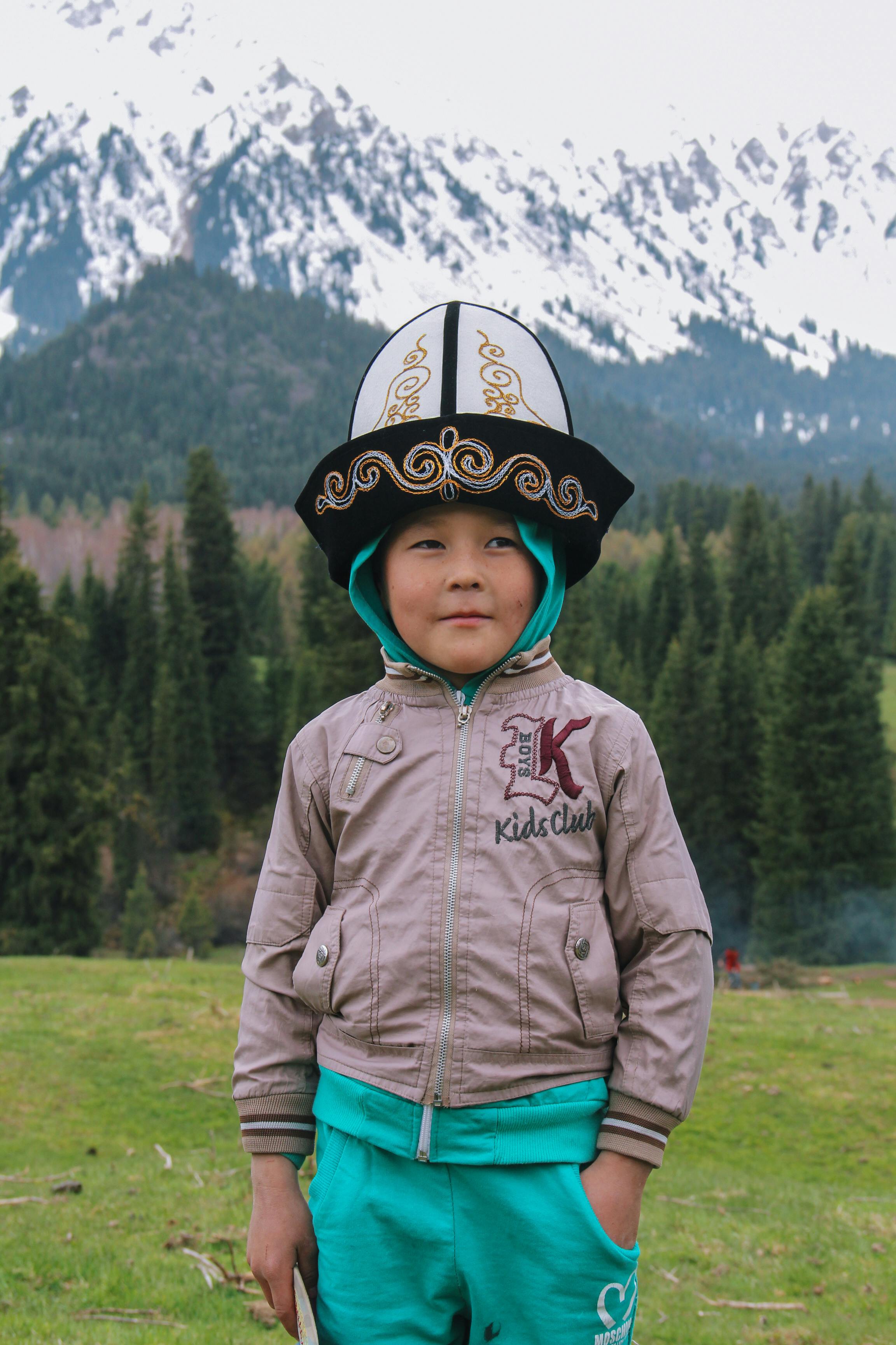 Young boy wearing a traditional hat stands before snow-capped mountains in Kyrgyzstan.