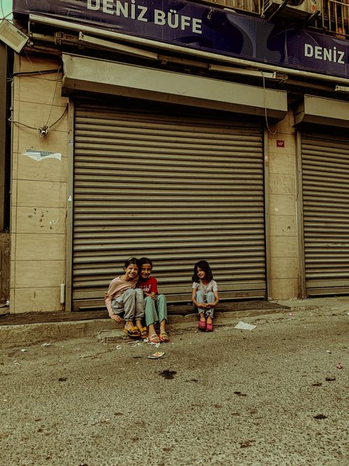 Free Three children sitting on a sidewalk by closed shop shutters on a city street. Stock Photo