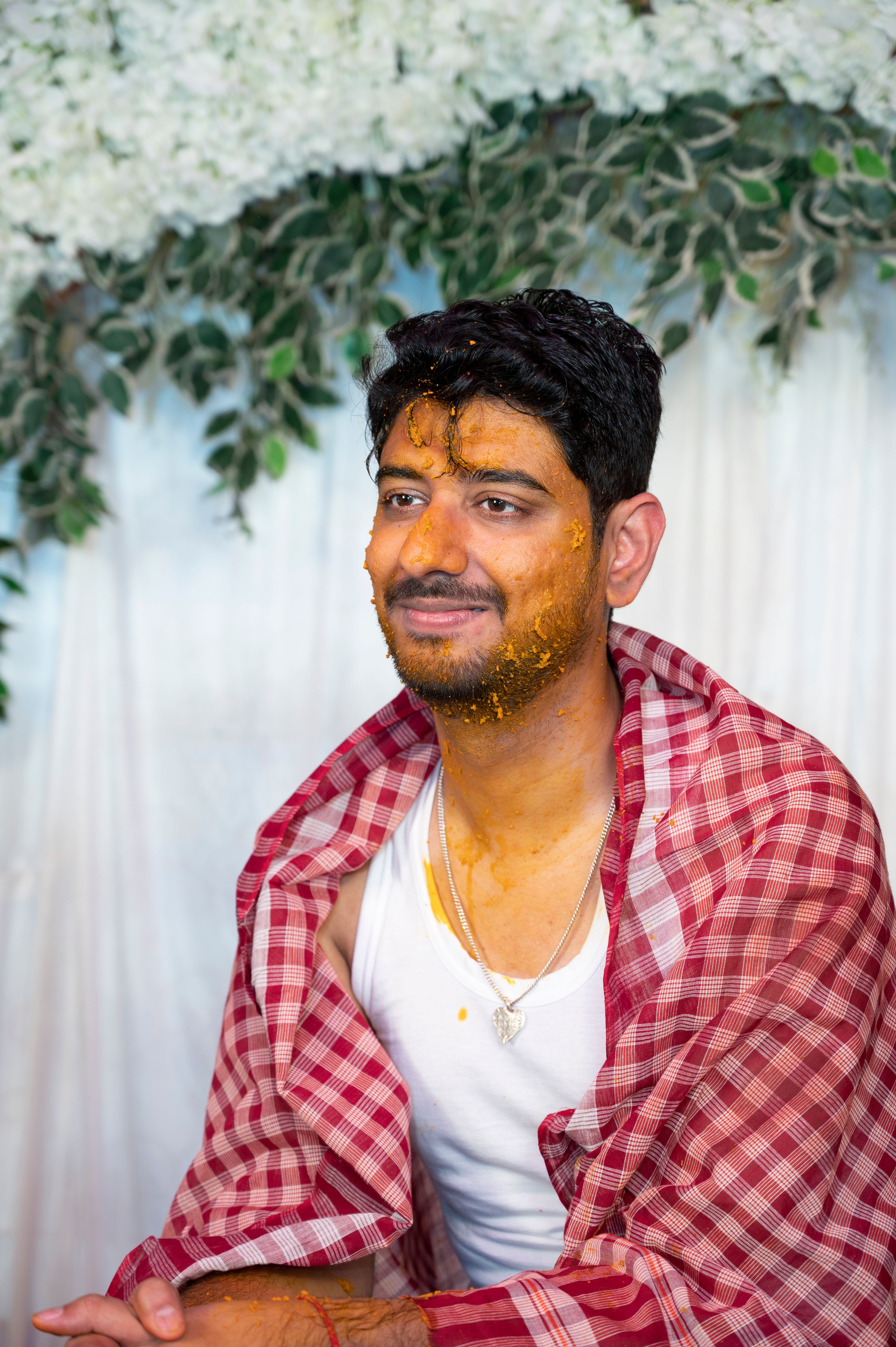 South Asian man covered in turmeric paste during a traditional Indian haldi ceremony with floral backdrop.