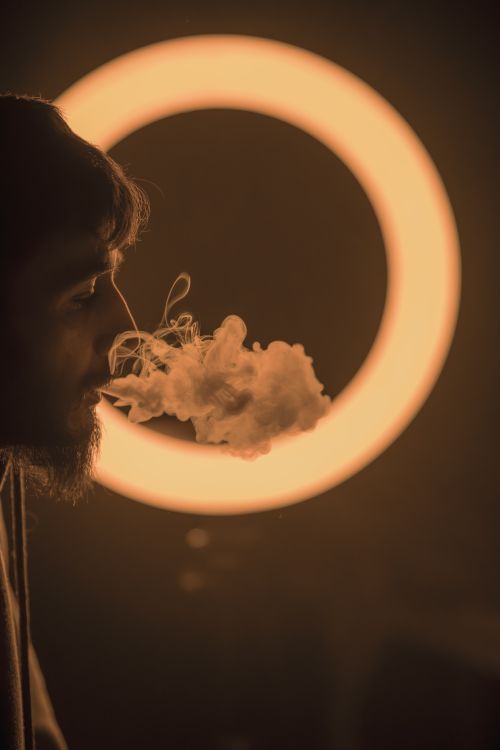 Free Creative portrait of a man exhaling smoke against a glowing circle backdrop, showcasing moody lighting. Stock Photo