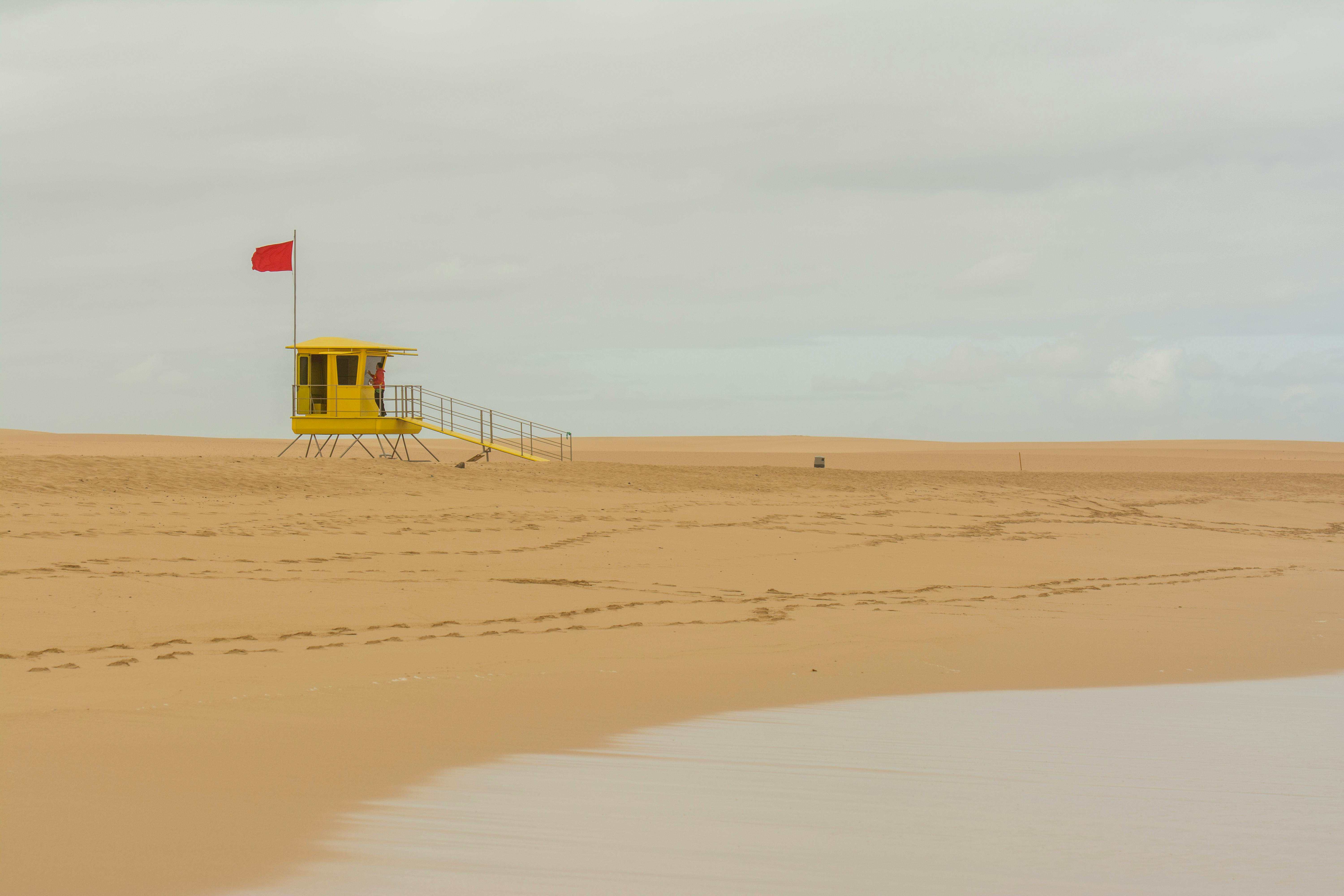 A yellow lifeguard tower with a red flag on a deserted sandy beach under overcast skies.
