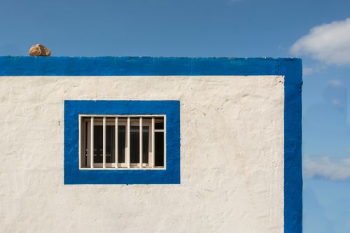 Free A minimalist white wall with a blue trim and barred window under a clear blue sky. Stock Photo