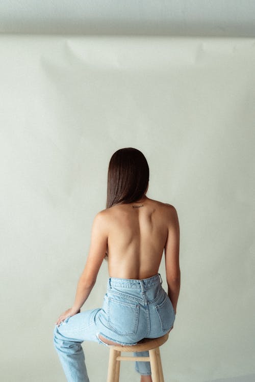 Topless Brunette Woman in Light Blue Jeans Sitting on a Stool