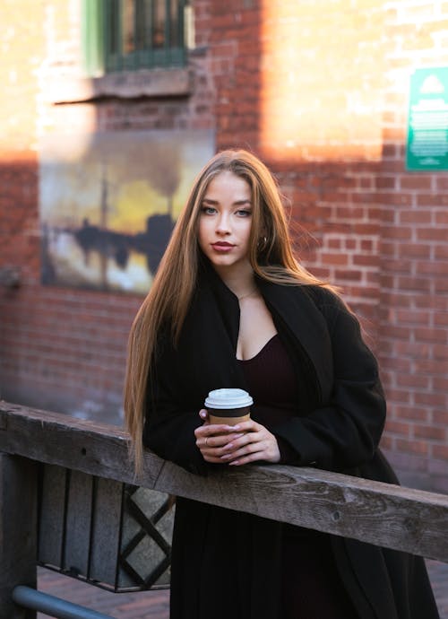 Model in a Long Black Coat and a Burgundy Dress Standing in an Alley by a Wooden Railing with a Cup of Coffee