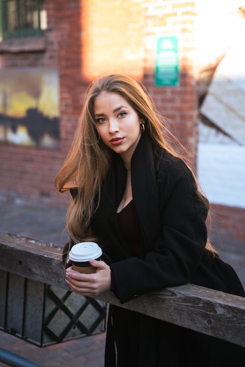 Model in a Long Black Coat in an Alley by a Wooden Railing with a Cup of Coffee
