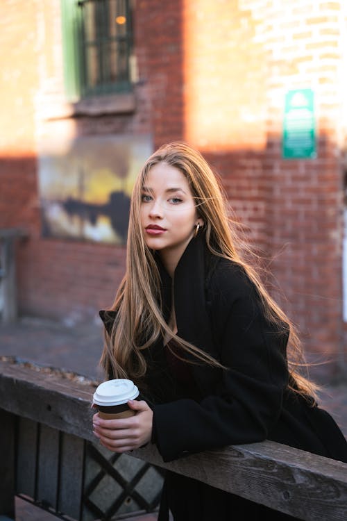 Young Woman in a Black Coat Standing by a Wooden Railing with a Cup of Coffee