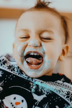Adorable toddler laughing with icing on face, wearing festive Christmas sweater indoors. Captures pure joy and fun.