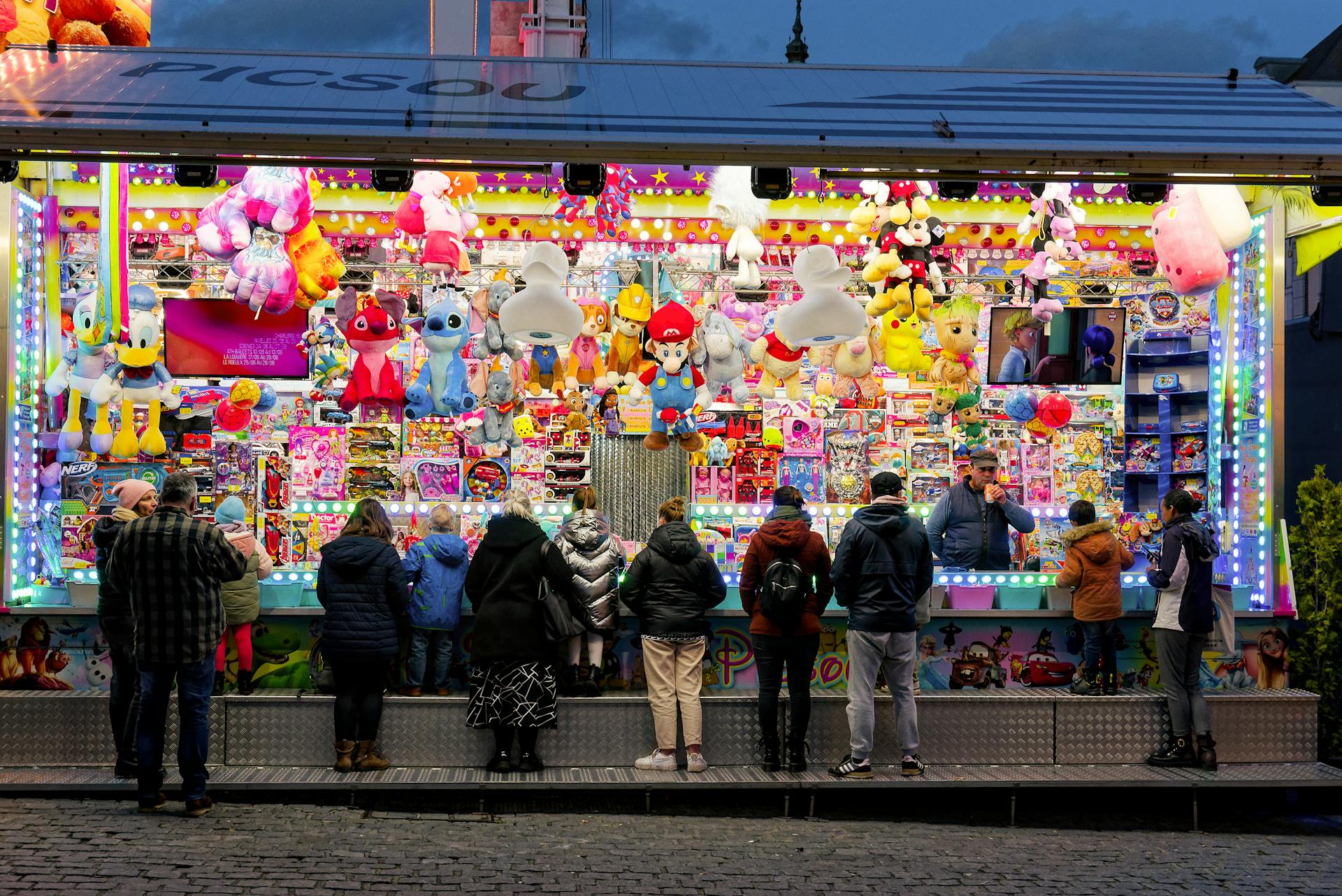 People at an illuminated carnival game stand with plush toys and vibrant lights at night.