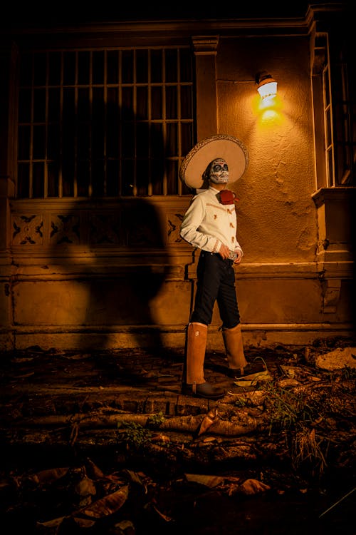 Man in Sombrero and with Painted Face Standing by Wall with Light at Night