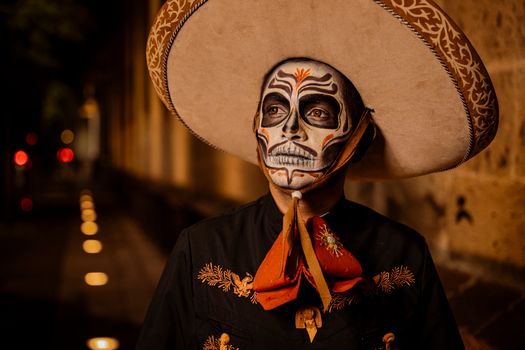 A man in Dia de los Muertos makeup and traditional attire stands outdoors at night wearing a sombrero.