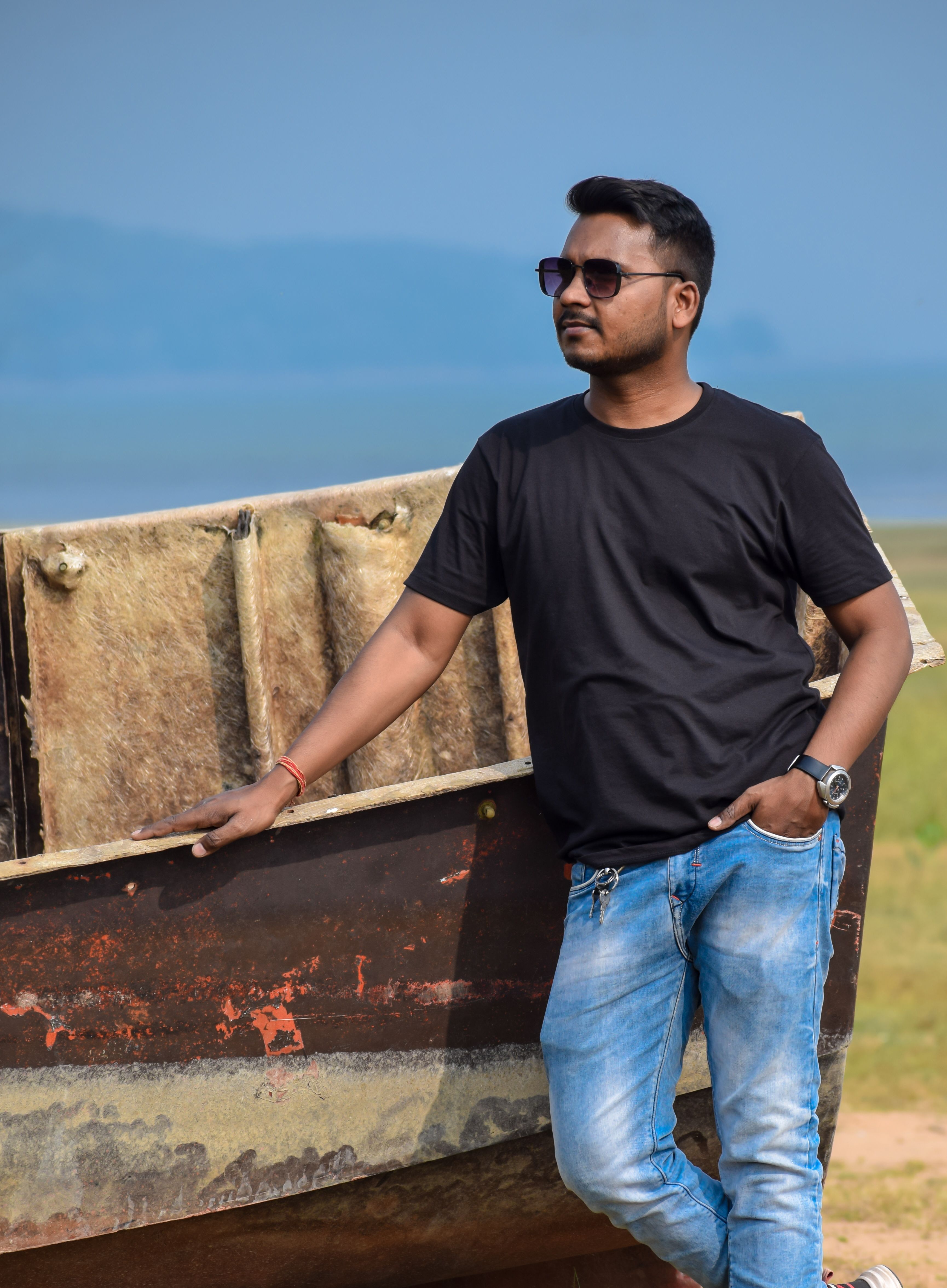 Man posing by a rustic boat in a countryside setting, under the summer sun.