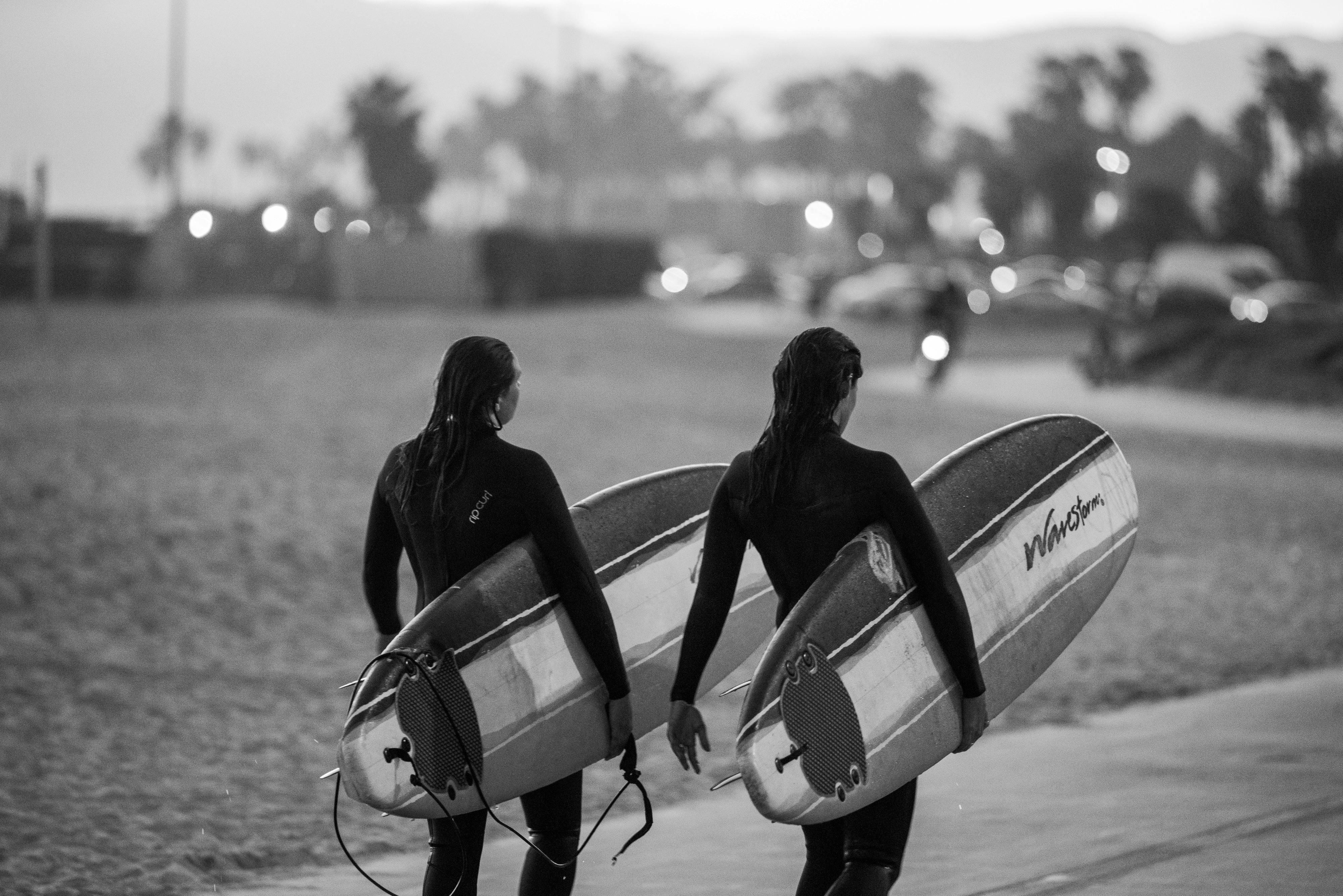Monochrome image depicting two surfers walking on a beach with surfboards in hand.