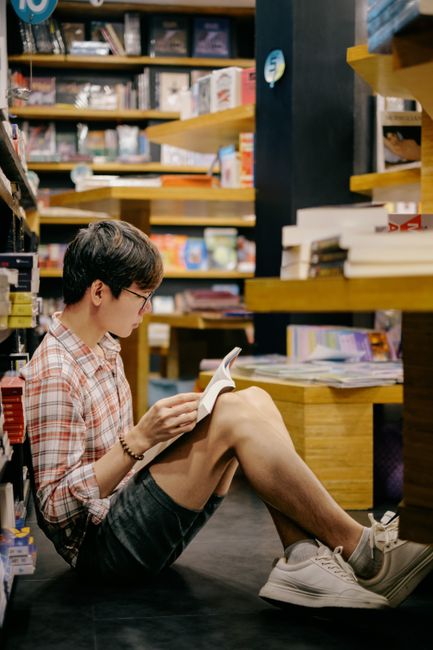 Casual young man reading a book while sitting on the floor of a bookstore, surrounded by shelves.