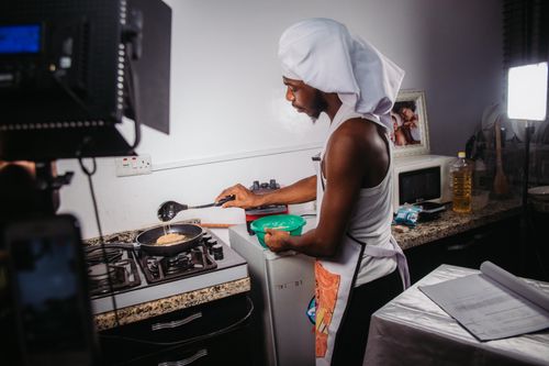 Free Man in apron and chef hat cooks in home kitchen with professional lighting setup. Stock Photo