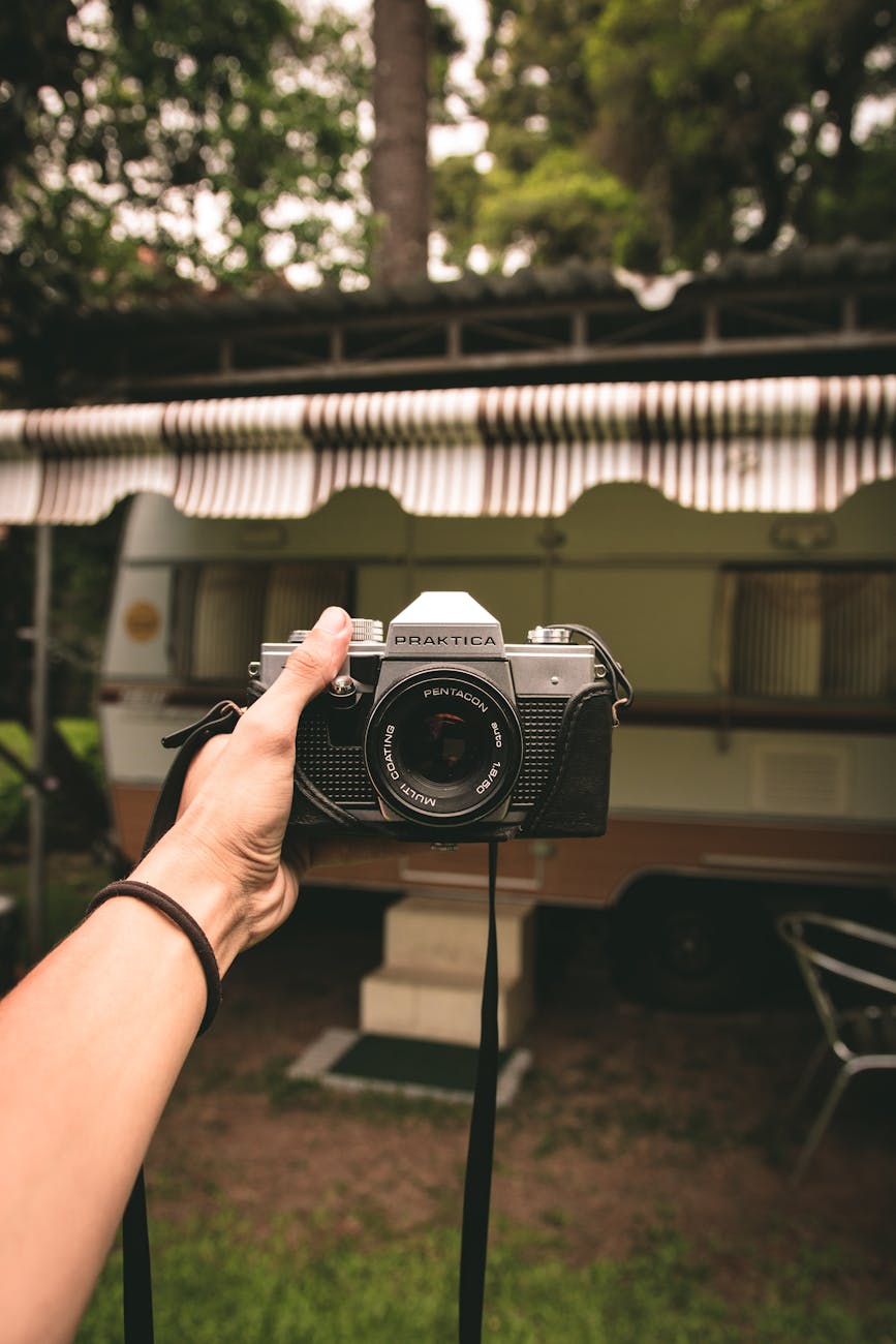 A hand holds a vintage camera with a campervan in the background, set in a lush outdoor campsite.